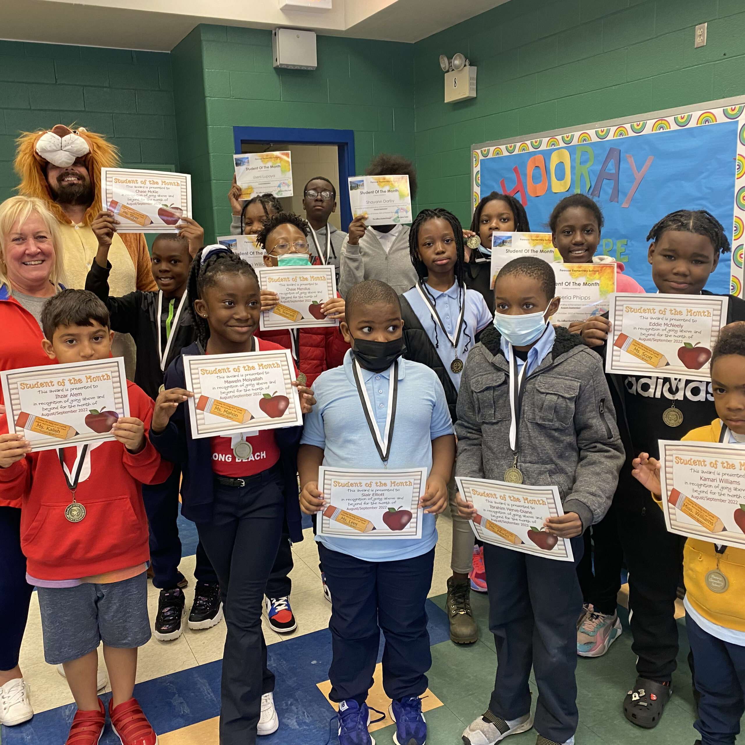 Student of the Month Celebration Penrose students holding Student of the Month certificates with medals, joined by staff and a person in a lion costume.